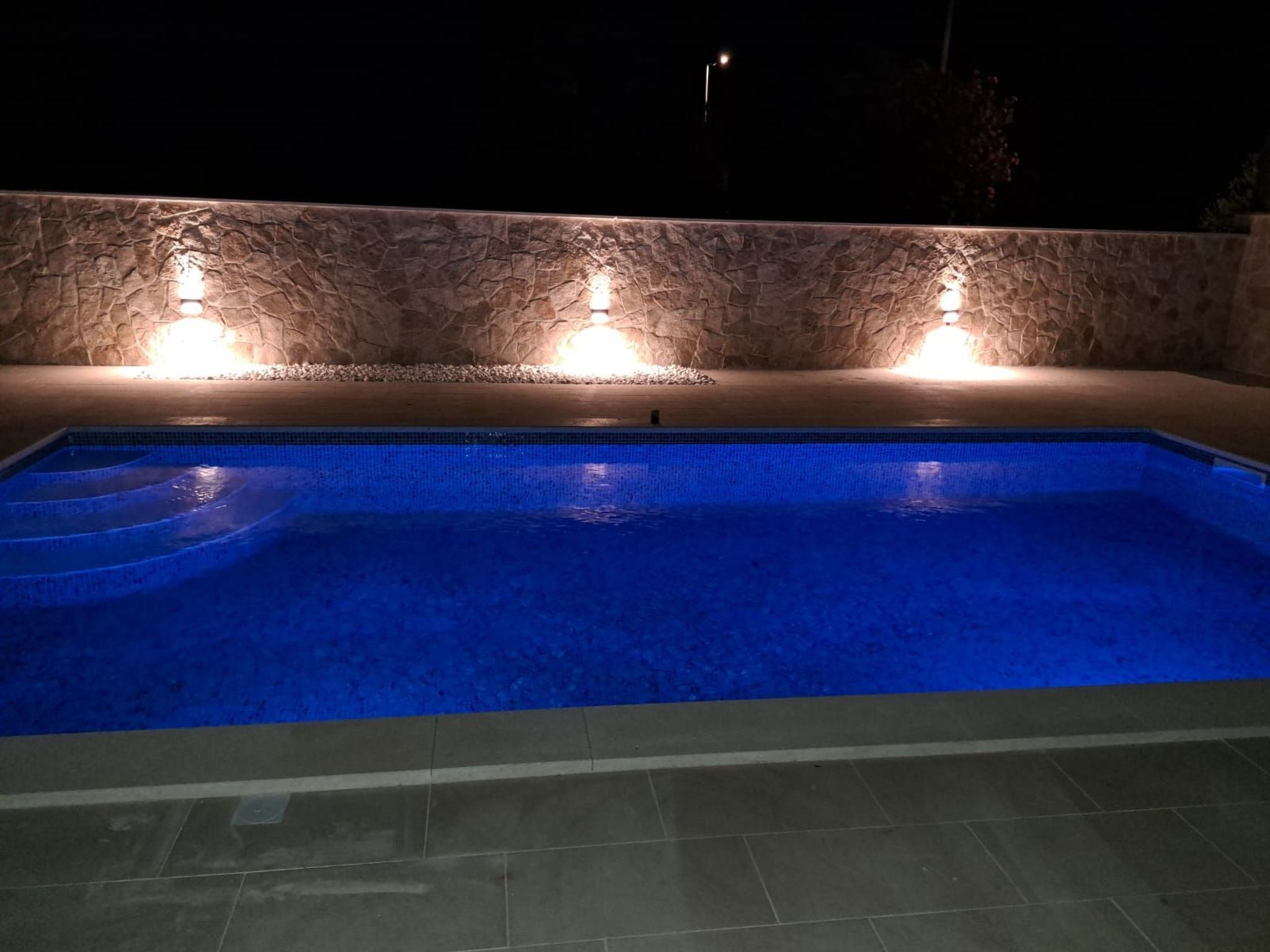 A swimming pool with blue tiles and a stone wall with lights.