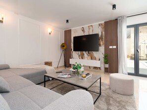 A modern living room with a large flat-screen TV mounted on a marble wall, grey sofas, a coffee table with magazines and a vase of flowers, and a sliding glass door leading to an outdoor area. - Designer Villa Simone - Modern und Heritage Style by Interhome (Jakovici)