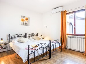 A bedroom with a metal bed frame, white bedding, a bedside lamp, a wall-mounted air conditioner, and a window with curtains.