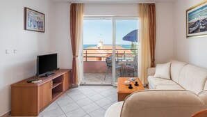 Quarto de hotel com televisão sobre um móvel de madeira, um sofá bege, uma mesinha com uma tigela e chávenas, e varanda com mesa e cadeiras.