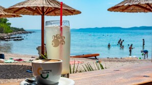 A beachside table with a glass of a clear beverage and a cup of coffee, with beach umbrellas and people in the background. - Mobilheim direkt am Strand by Interhome (Ugljan/Ugljan)