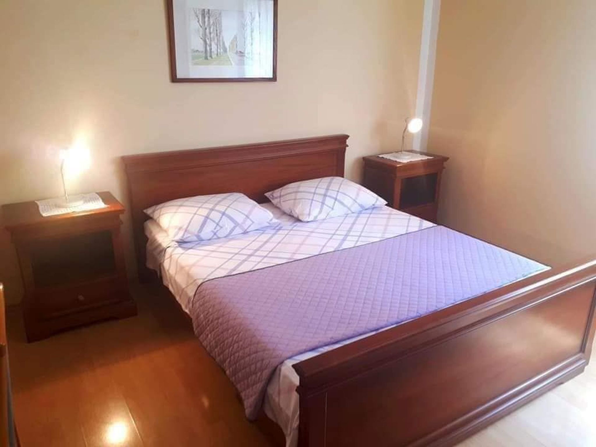 Een netjes opgemaakt bed met een geruit sprei, twee nachtkastjes met lampen, en een ingelijst beeld aan de muur.