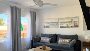 Moderni olohuone, jossa on harmaa sohva, kaksi koristetyynyä, joissa on teksti ”BEACH”, kattotuuletin ja taideteoksia, joissa on kuvattuja rantamaisemia.