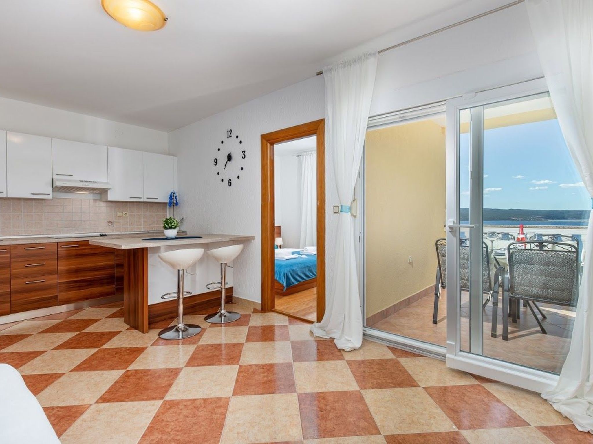 A modern kitchen with a checkered floor, a dining area with bar stools, a bed with a blue bedspread, and a balcony with outdoor furniture.