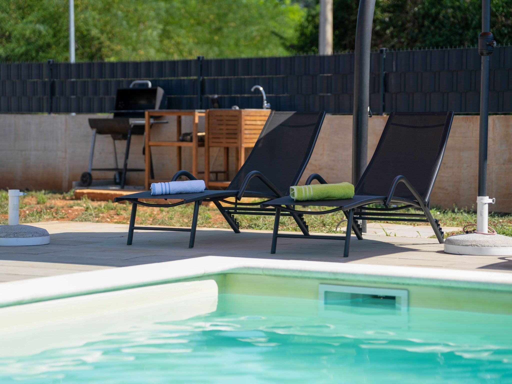 Un espace aménagé au bord de la piscine, avec deux chaises longues, un barbecue et un mur en béton en arrière-plan.