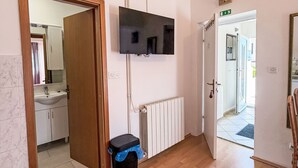 A hallway with a wooden door, a black trash bin, a white radiator, and a mounted television. - Studioapartment Meri 4 by Interhome (Losinj / Mali Losinj)