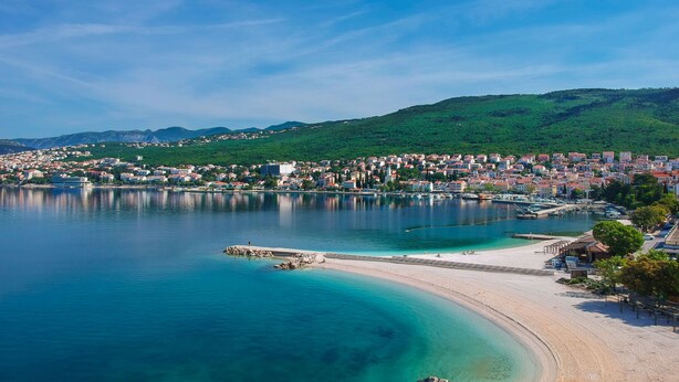 Uma cidade costeira com praia de areia, águas azul-claras e uma área densamente construída, com edifícios e colinas ao fundo.