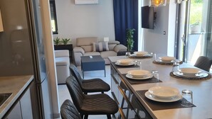 Ein moderner Essbereich mit einem langen Tisch, gedeckt für vier Personen, einer Couch im Hintergrund und einer sichtbaren Küchenzeile links.