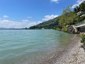 Um lago calmo com uma praia de pedras e uma colina com edifícios.