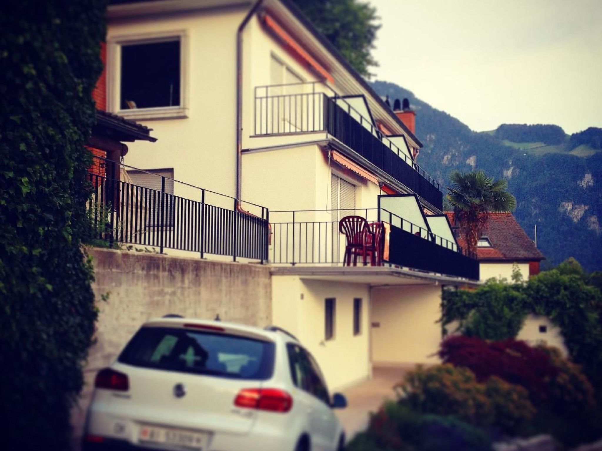 Una casa de dos pisos con un balcón, un coche estacionado adelante y montañas al fondo.