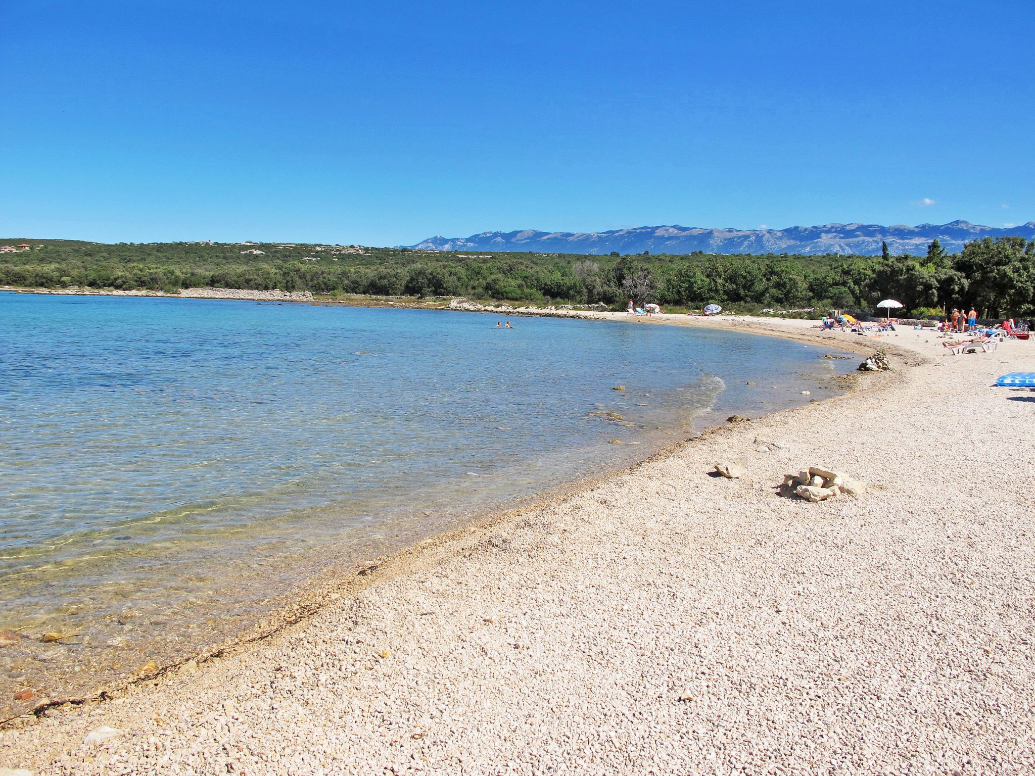 Une plage de galets avec une eau claire, quelques personnes et des montagnes en arrière-plan.