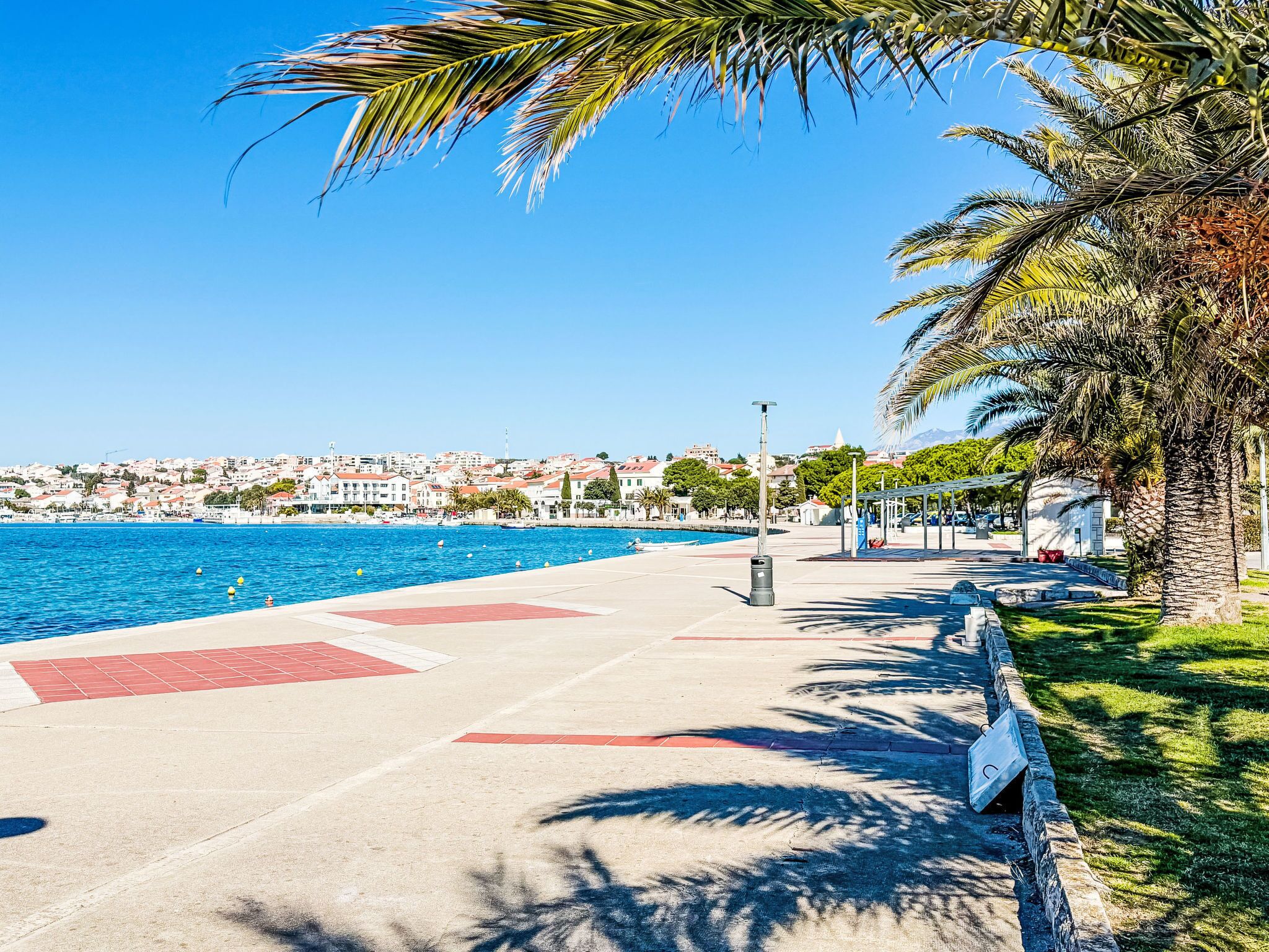 A coastal promenade with a clear blue sea, a paved walkway, palm trees, and a distant town.