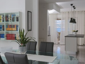 Uma sala de estar moderna com uma mesa de jantar de vidro, cadeiras pretas, uma estante, uma planta em vaso e vista para o mar através de cortinas leves.