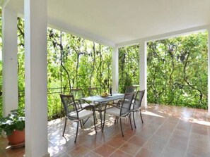Eine überdachte Terrasse mit Tisch und Stühlen, umgeben von weißen Säulen und Blick auf üppiges Grün.