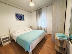 Ein Schlafzimmer mit Bett, Nachttisch, einem an der Wand hängenden Bild, einem Fenster mit Vorhängen und Holzboden.
