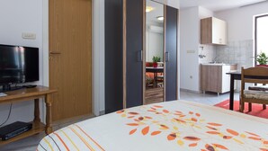 Ein modernes Zimmer mit einem Bett, einem Fernseher, einem Kleiderschrank, einem Spiegel, einem Schreibtisch und einer Küchenzeile.