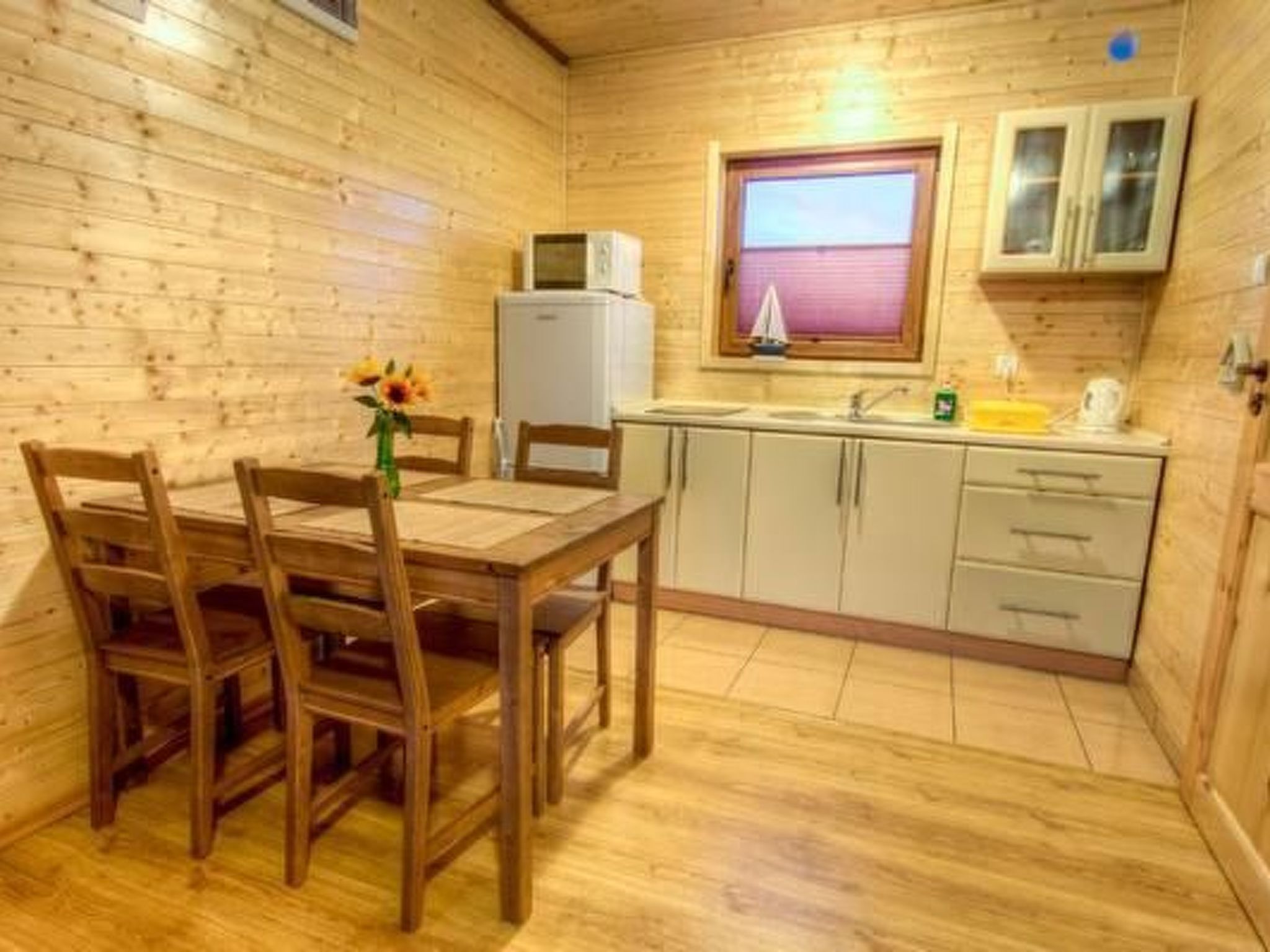 A wooden cabin interior with a dining table, chairs, a kitchen area including a sink and cabinets, and a window with blinds.