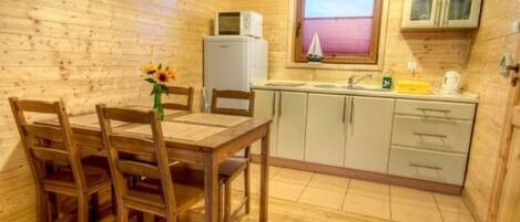 A wooden cabin interior with a dining table, chairs, a kitchen area including a sink and cabinets, and a window with blinds.