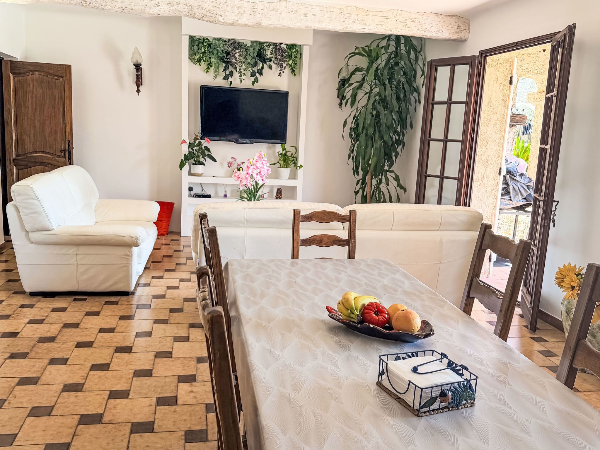 Una sala de estar con un sofá, una mesa de comedor con fruteros, un televisor montado en la pared y una planta grande.