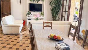 Una sala de estar con un sofá, una mesa de comedor con fruteros, un televisor montado en la pared y una planta grande.