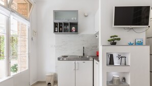 Een compacte keuken met een ingebouwde kookplaat, witte kastjes en een marmeren spatwand. Een kleine prullenbak is zichtbaar bij de ingang, en een televisie hangt aan de muur boven een plank met decoratieve objecten.