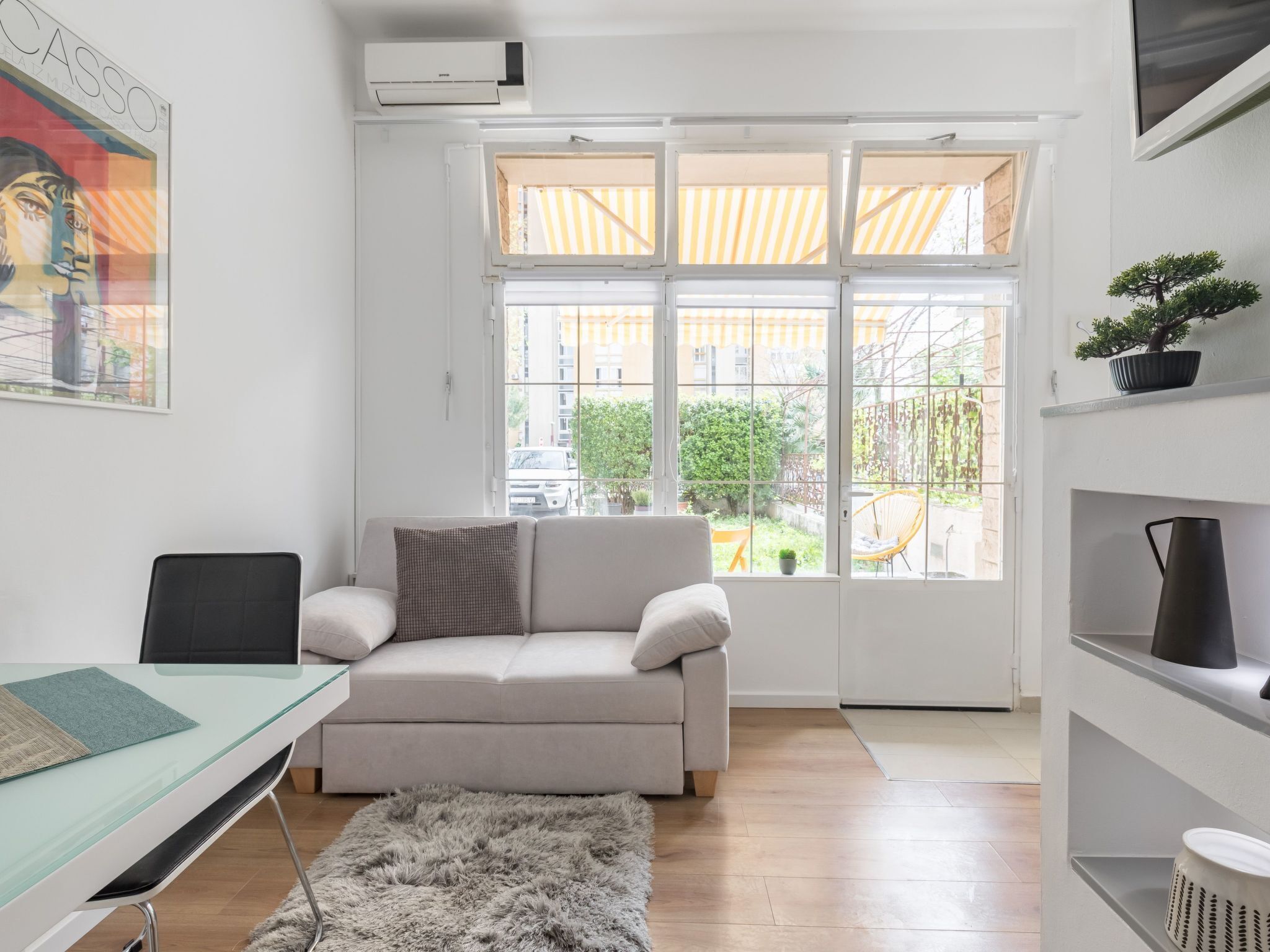 A modern living room with a sofa, a glass desk, a fluffy rug, and a large window with a view of a courtyard and buildings.