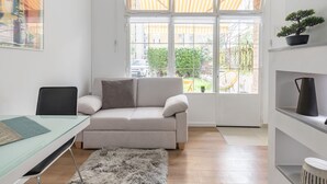 Een moderne woonkamer met een bank, een glazen bureau, een zacht vloerkleed en een groot raam met uitzicht op een binnenplaats en gebouwen.