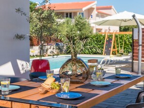 Uma área de refeições ao ar livre com piscina ao fundo, mesa de madeira com pratos, copos e um vaso com folhagem.
