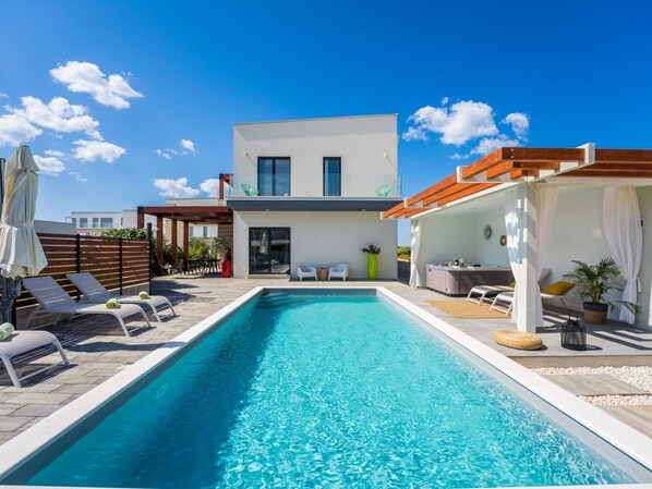 Uma área de piscina moderna com piscina retangular, espreguiçadeiras e um espaço coberto com mesa e cadeiras.