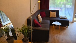 A modern living room with a dark grey sofa, red and beige cushions, a small white side table, and a tall floor lamp. There are two potted plants on the floor. - Haus Ahorni by Interhome (Beatenberg)