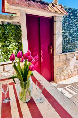 Ein Tisch mit einer Vase voller pinkfarbener Tulpen und zwei leeren Weingläsern, vor einer roten Tür und einer Steinwand.