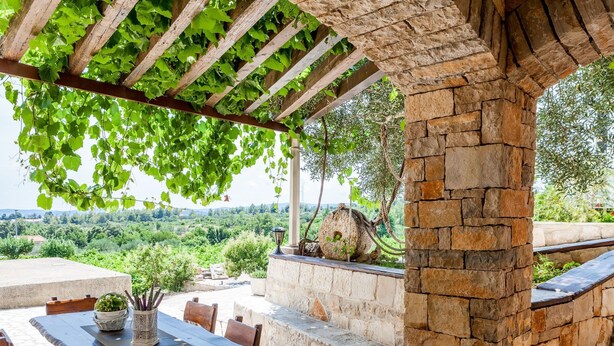 Un área de comedor al aire libre con una mesa y sillas de piedra, una pérgola con enredaderas verdes y un muro de piedra con un objeto decorativo grande.