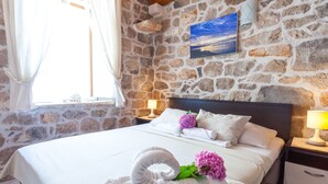 Un dormitorio con pared de piedra, una cama con sábanas blancas, un cuadro y una ventana con cortinas.