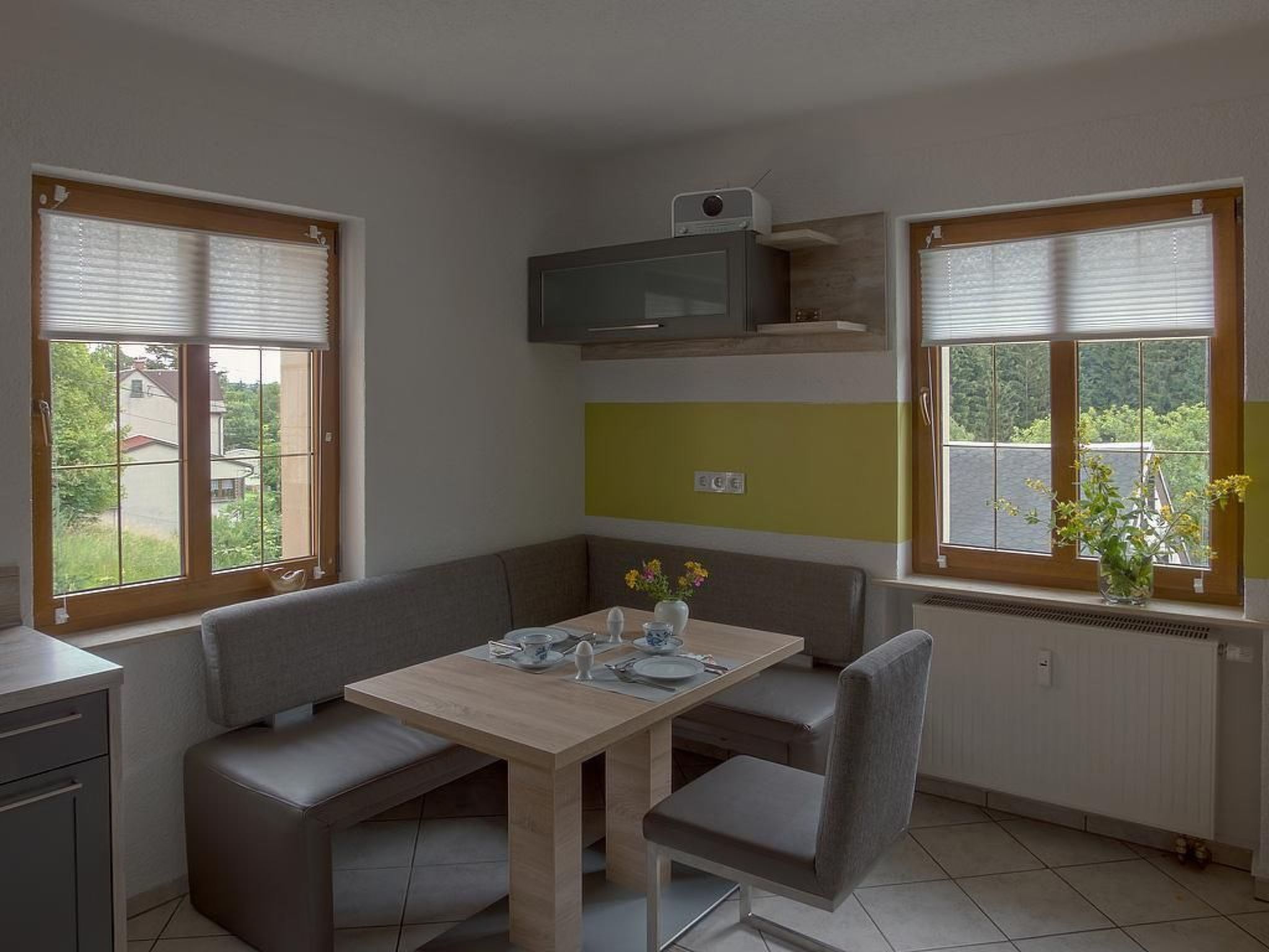 A dining area with a table, chairs, and a bench. There are two windows with blinds, a microwave above the table, and a radiator below the windows.