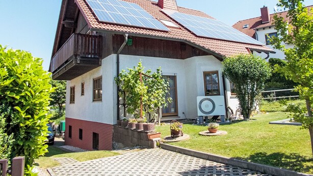 Ein zweigeschossiges Haus mit rotem Ziegeldach, Solaranlage, einem Balkon und einem Garten mit Kübelpflanzen.