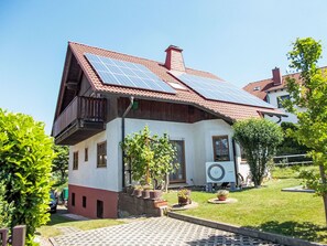 Une maison à deux étages avec un toit recouvert de tuiles rouges, des panneaux solaires, un balcon et un jardin agrémenté de plantes en pot.