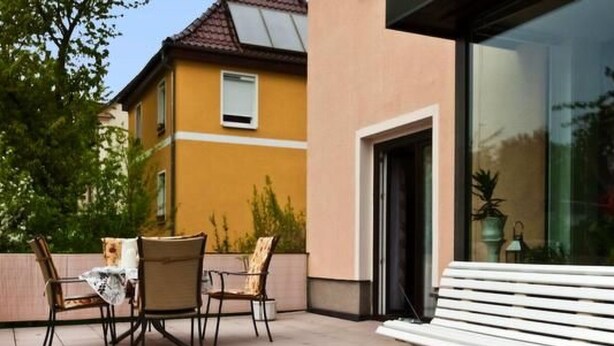 Une terrasse extérieure avec un banc blanc et deux chaises, une porte vitrée donnant sur un bâtiment, et un mur jaune en arrière-plan.