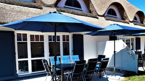 Een terras buiten met een tafel en stoelen, overdekt met blauwe parasols, en een wit gebouw met rieten dak op de achtergrond.