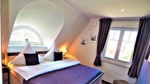Een slaapkamer met een bed, een nachtkastje, een lamp, een raam met gordijnen en een klein krukje.