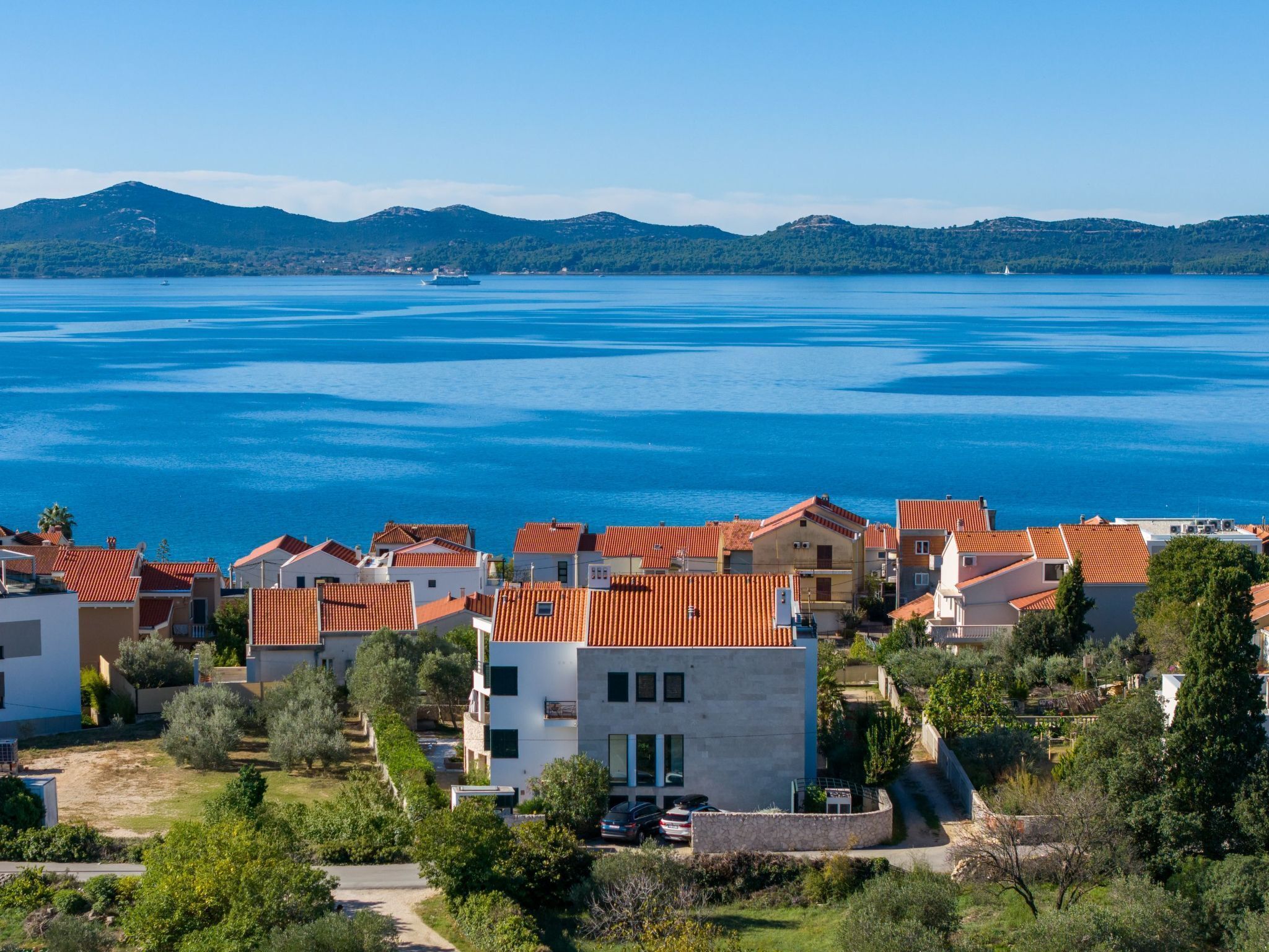 Une zone résidentielle côtière avec des maisons aux toits de tuiles rouges, une mer d’un bleu limpide et des montagnes à l’horizon.