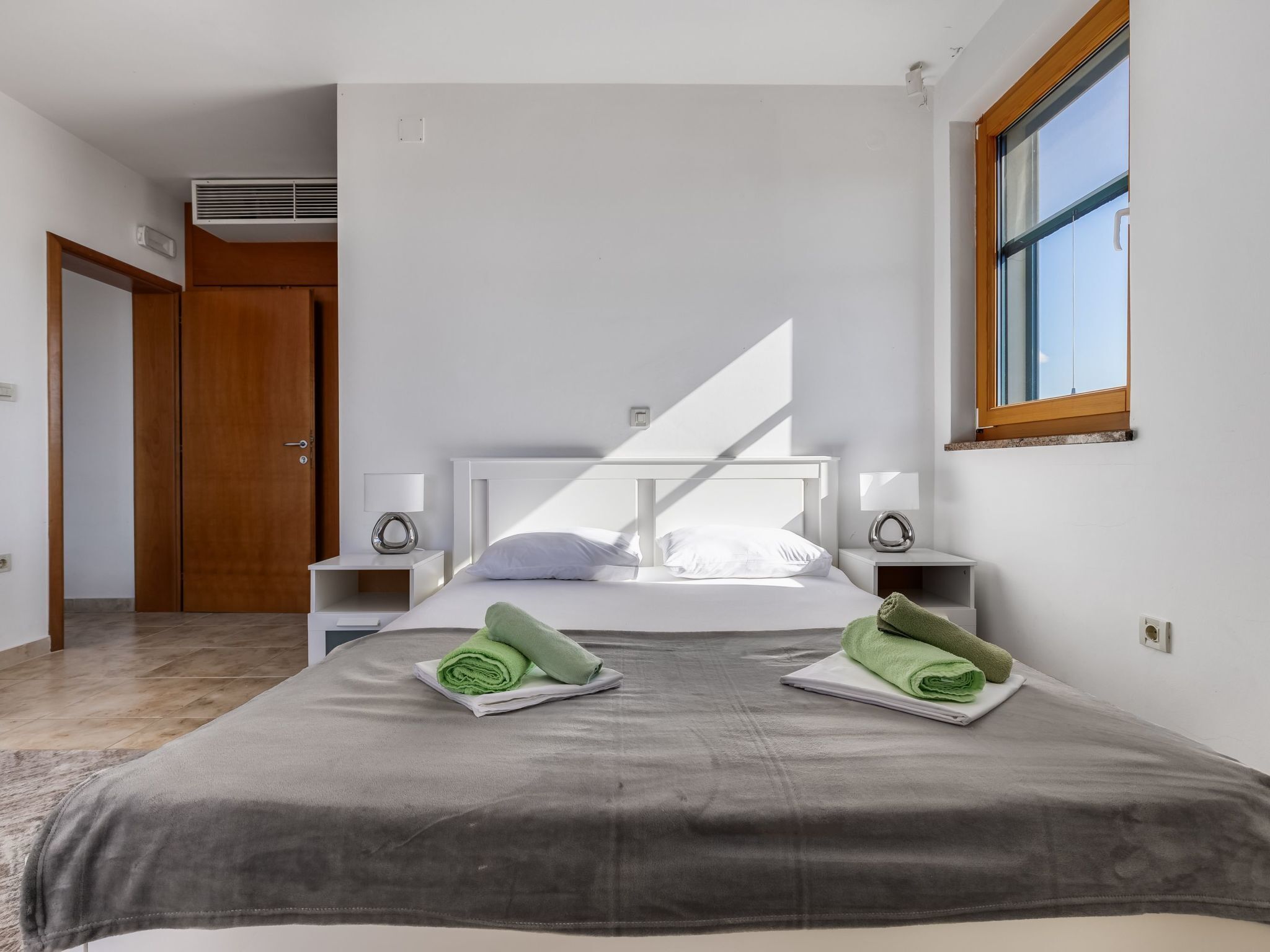A modern hotel room with a large bed, two bedside tables, a window, and a door.