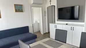 Uma sala de estar moderna com um sofá azul, uma mesa de jantar com jogos americanos cinzas, armários brancos e uma TV na parede.