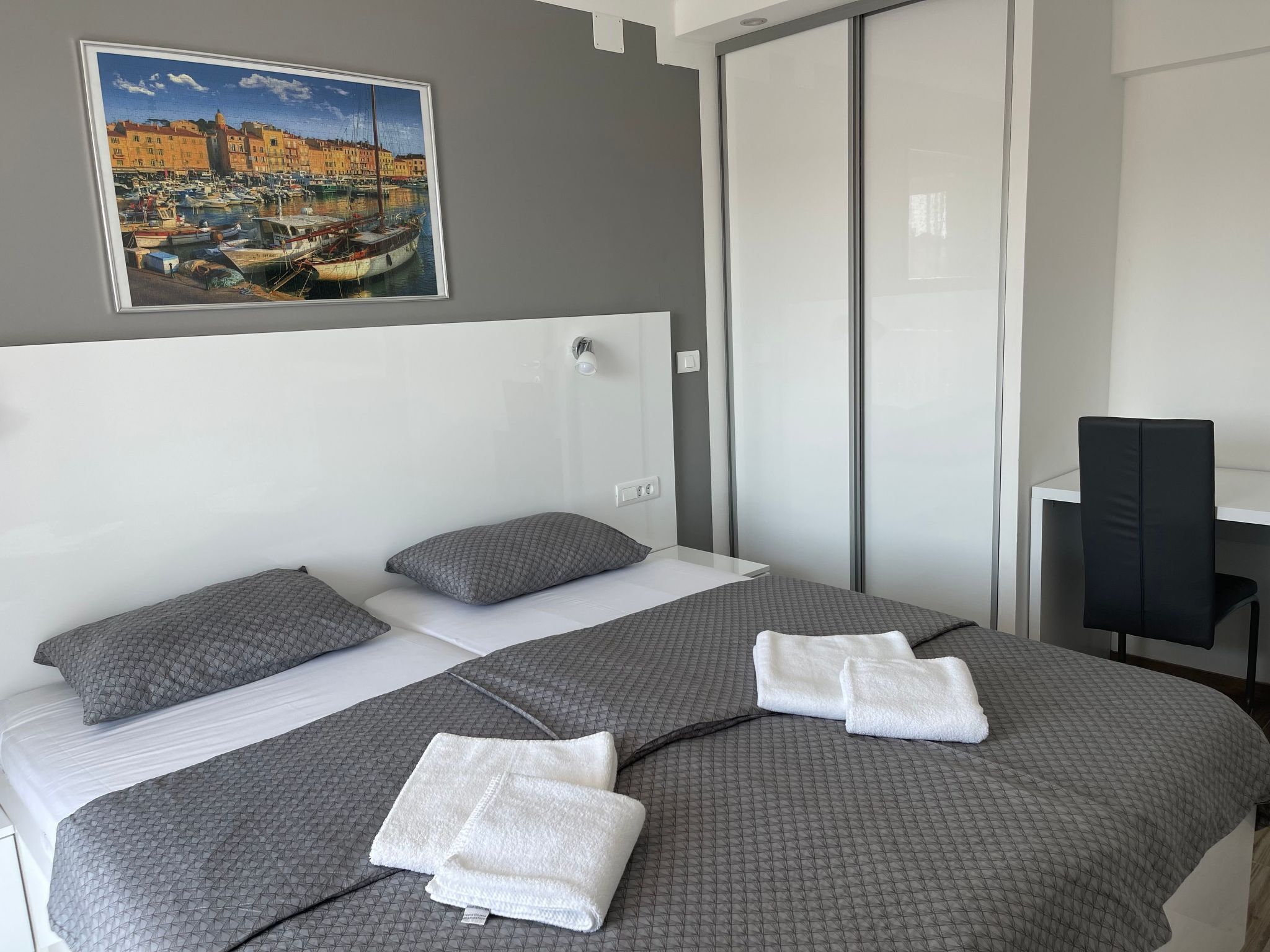 A hotel room with a bed, two pillows, a white headboard, a painting of a harbor, and a desk with a chair.