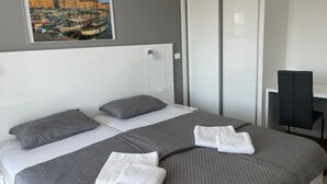 Quarto de hotel com cama, duas almofadas, cabeceira branca, pintura de um porto e escrivaninha com cadeira.
