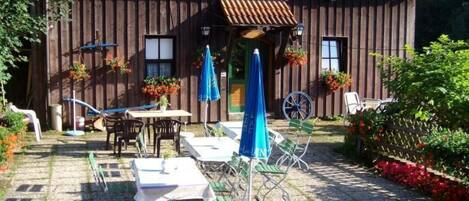 Et træhus med tegltag, udendørs siddeområde med borde og stole, samt en have med blomster.