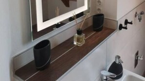 Un baño con un espejo grande, un tocador de madera, un bote de basura negro, una botella pequeña de perfume y una bañera con un tapete negro.