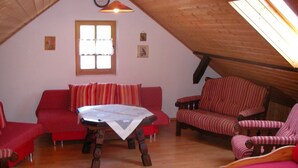Een gezellige zolderkamer met een schuine kap, houten vloeren, een rode bank, een gestreepte fauteuil en een klein tafeltje met een witte tafelkleed.