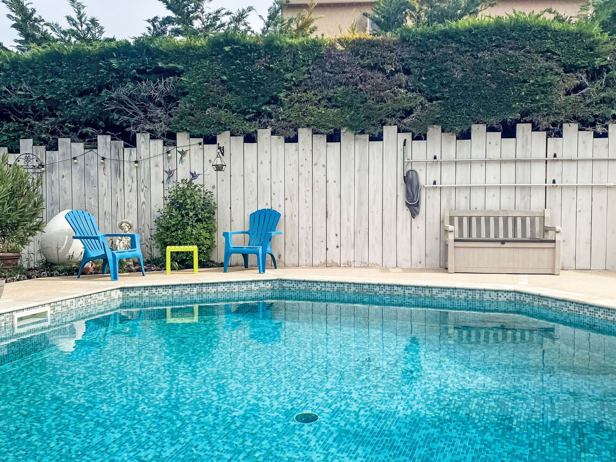 A swimming pool with blue chairs and a small table, surrounded by a wooden fence and greenery.