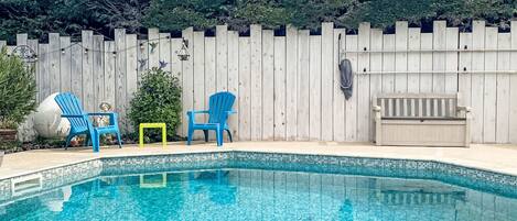 A swimming pool with blue chairs and a small table, surrounded by a wooden fence and greenery.