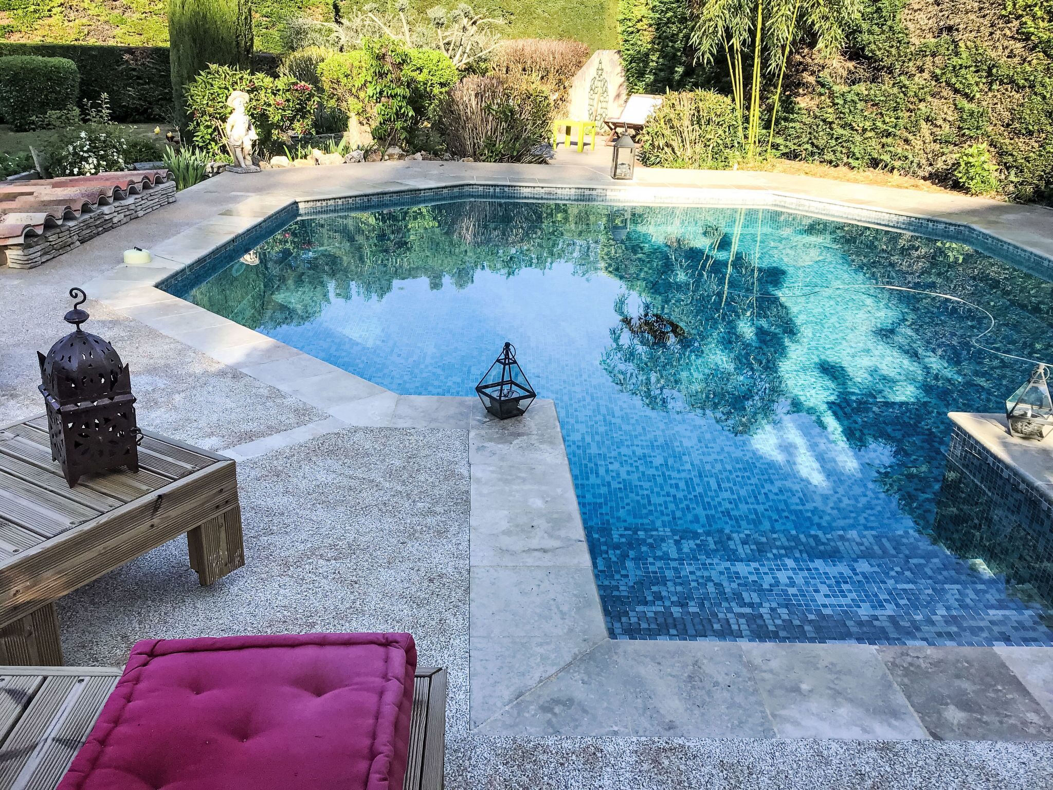 Une piscine dont le bord est revêtu de carreaux, entourée d’un jardin aménagé avec différentes plantes et une statue.